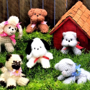 May include: A collection of crocheted animal toys, including a poodle, a brown dog, a white dog, a Siamese cat, and a gray cat. The toys are sitting on a bed of green grass with a red doghouse in the background.