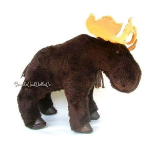 Pode incluir: Um brinquedo de pelúcia de alce marrom com uma grande galhada de feltro laranja. O alce tem um focinho longo e olhos pretos. O brinquedo está em pé sobre uma superfície branca.