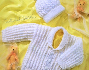 May include: White knitted baby cardigan with a button closure and a matching white knitted hat with a pom pom. The cardigan and hat are laid out on a yellow satin fabric.
