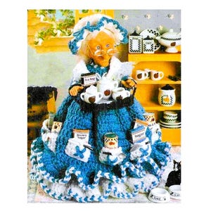 Puede incluir: Una muñeca de crochet con un vestido azul y un delantal blanco, sosteniendo una bandeja con tazas de té y una tetera. La muñeca está rodeada de varios artículos de cocina, incluyendo botes, cuencos y un gato.