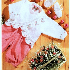 May include: A white knitted baby outfit with pink accents, including a long-sleeved top with floral embroidery and a scalloped collar, paired with pink corduroy pants and white baby shoes. The scene is decorated with roses.