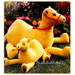 Puede incluir: Dos juguetes de peluche de camello, uno grande y otro pequeño, están tumbados en un lecho de hierba verde. El camello más grande lleva una brida roja. Ambos camellos son de color marrón claro.