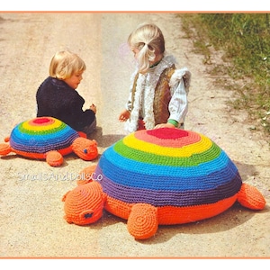May include: Two large, colorful, crocheted turtle-shaped pillows. The turtle shells feature a rainbow pattern, and the bodies are orange. The pillows are on the ground with two children nearby. The text "SmallsandDollsCo" is visible.