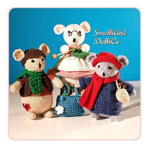 Op de afbeelding: Drie handgebreide muispoppen met de tekst SmallsAnd DollsCo. De linker muis draagt een bruin jasje en een groene sjaal. De middelste muis draagt een roze jurk en houdt breinaalden vast. De rechter muis draagt een blauwe trui en een rode sjaal.