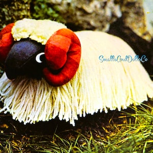 May include: A white fluffy sheep toy with red horns and a black face. The toy is lying on a bed of green grass.