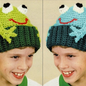 Könnte beinhalten: Zwei Kinder tragen grüne und blaue Häkelmützen mit Frosch-Motiv. Die Mützen haben weiße Augen, schwarze Pupillen und rote lächelnde Münder.