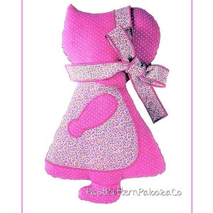 May include: A pink fabric doll in the shape of a girl, with a bonnet and apron. The bonnet and apron feature a floral pattern. The doll has a large bow and is made of pink polka dot fabric. The text "PastPatternPaloozaCo" is at the bottom.