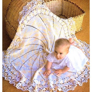 May include: A white crocheted baby blanket with a delicate lace border. The blanket is laid out on a brown carpet with a wicker basket in the background. A baby is lying on the blanket.