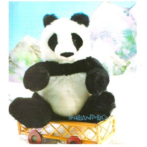 May include: A black and white panda bear plush toy sitting in a small wooden wagon with red wheels. The panda is wearing a black band around its waist. The wagon is on a snowy surface with a blue sky and white mountains in the background. The text "SmallsAndDollsCo" is printed on the wagon.