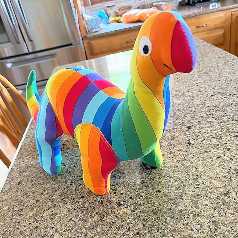 Puede incluir: Un juguete de dinosaurio de peluche a rayas de arco&iacute;ris. El dinosaurio est&aacute; de pie sobre una superficie marr&oacute;n claro.