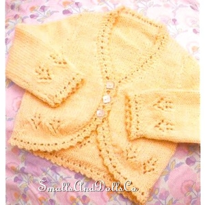 May include: A pale yellow knitted cardigan with a scalloped edge and leaf-shaped cutouts on the sleeves. The cardigan has three small, square buttons and the text "SmallsAndDollsCo" is visible. The garment is displayed on a floral patterned fabric.