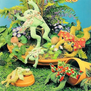 Puede incluir: Cuatro peluches de rana verde con lazos de tela de diferentes patrones. Las ranas están colocadas en un entorno de jardín con hojas verdes y macetas marrones.