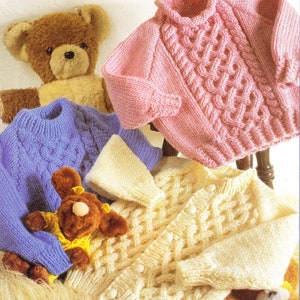 Op de afbeelding: Drie gebreide babytruien in roze, blauw en crème met een kabelbreipatroon. De truien worden getoond op een stoel met knuffels.