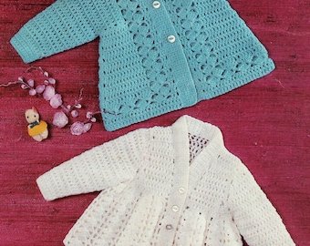May include: Two crocheted baby jackets, one in light blue and one in white, laid out on a red surface. Both jackets have a button closure and a lacy design.