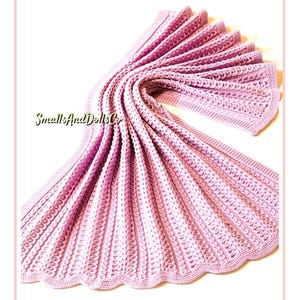 May include: A light pink, knitted blanket with a scalloped edge. The blanket is folded and draped, showcasing its textured pattern. The text "SmallsAndDollsCo" is visible in the lower left corner. The blanket appears soft and cozy.
