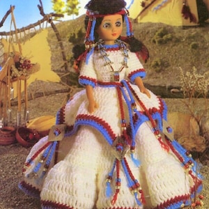 May include: A crocheted doll wearing a white dress with blue and red trim, a beaded necklace, and a feathered headdress. The doll is standing in front of a teepee.