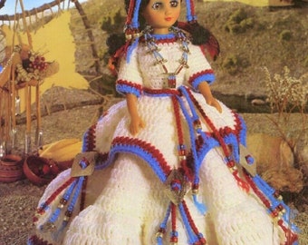 May include: A crocheted doll wearing a white dress with blue and red trim, a beaded necklace, and a feathered headdress. The doll is standing in front of a teepee.