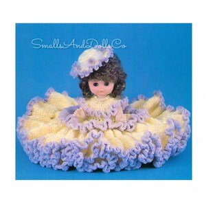May include: A crocheted doll dress with a yellow bodice and purple trim. The doll is wearing a purple and white crocheted hat. The doll has brown hair and brown eyes. The text "Smalls And Dolls Co" is visible in the top left corner of the image.