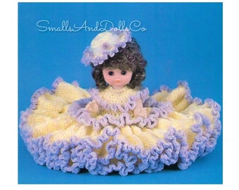 May include: A crocheted doll dress with a yellow bodice and purple trim. The doll is wearing a purple and white crocheted hat. The doll has brown hair and brown eyes. The text "Smalls And Dolls Co" is visible in the top left corner of the image.