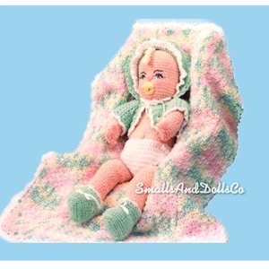 May include: A crocheted baby doll wearing a green and white outfit and a pink and white crocheted blanket. The doll is holding a pacifier and has a pink and white crocheted hat. The blanket has a green, pink, and white pattern.