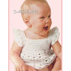 May include: A vintage image of a baby wearing a white, crocheted top with short sleeves. The top has a square neckline and a delicate, openwork design. The baby is smiling with their mouth open. The image has a pink border and the text "SmallCo and Dolls Co".