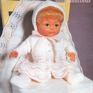 Puede incluir: Un conjunto de muñeca de bebé de punto blanco con ribete rosa y un gorro a juego. La muñeca está sentada en una manta de punto blanco.