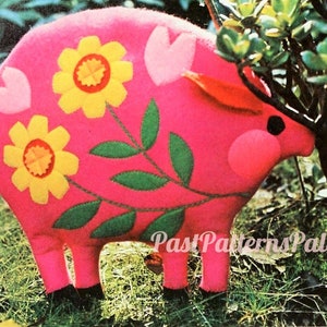 May include: A pink felt pig with a curly tail, green leaves, and yellow flowers sewn on. The pig is standing in a grassy area.