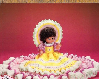 May include: A crocheted doll bed with a yellow, pink, and green ruffled design. A doll with brown hair and a yellow dress is sitting on the bed. The doll is wearing a pink and green crocheted hat. The image includes the text "Bed Dolls & Sweet Dreams" and "Free Gift Tags".