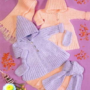 Puede incluir: Dos conjuntos de punto para muñecas, uno en melocotón y otro en lavanda, con gorros y bufandas a juego. Los conjuntos están dispuestos sobre un fondo morado con pétalos de flores secas.