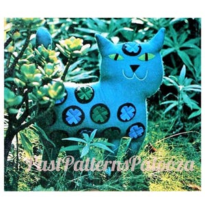 May include: A blue felt cat with black and green circles on its body. The cat is sitting in a green grassy area.