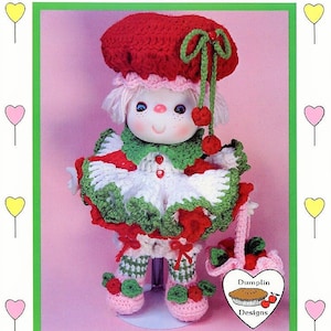 May include: A crochet doll with a red and white dress, green hat, and a red and white heart-shaped purse. The doll is standing on a white stand. The doll is labeled "Cherries Jubilee" and "Dumplin Designs".