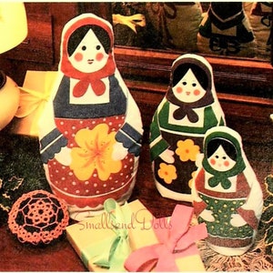 May include: Three nesting dolls, also known as Russian dolls, are made of fabric and feature a floral pattern. The largest doll is red with yellow flowers, the middle doll is green with yellow flowers, and the smallest doll is green with white polka dots. The dolls are sitting on a wooden surface with a gift box and a crocheted doily.