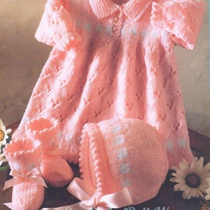 May include: A pink knitted baby dress, hat and booties set with white accents. The dress has a collar and a button fastening. The hat has a scalloped edge. The booties have bows.