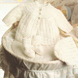 Puede incluir: Un conjunto de ropa de bebé de ganchillo blanco que incluye un cárdigan, botines y un gorro.
