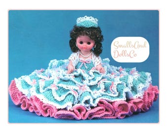May include: A doll with dark curly hair wearing a crocheted teal and white dress with pink ribbon accents, a white bolero, and a matching teal crown. The doll features a pearl necklace and the text SmallsAnd DollsCo is visible in the corner.