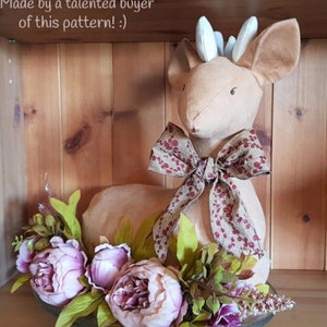 May include: A beige fabric deer with a red and brown bow, sitting on a glass plate with pink peonies and green leaves. The deer has white antlers and black eyes. The text "Made by a talented buyer of this pattern! :)" is visible above the deer.