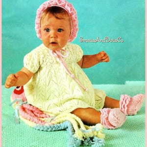 以下が含まれることがあります： ピンクの編み物製の帽子とそれに合わせたピンクの靴下を履いた赤ちゃんが、黄色のニットのドレスを着ています。赤ちゃんはカラフルな編み物製の玩具に座っています。画像には「Smallso And Dolls Co.」というテキストが含まれています。