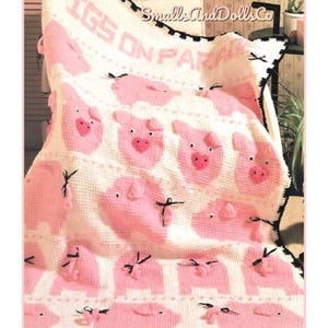 以下が含まれることがあります： 「PIGS ON PARADE」の文字とピンク色の豚の模様が繰り返される、白とピンクの手編みのブランケット。ブランケットには黒い縁取りと小さな黒いリボンが付いています。手作りの魅力的なアイテムです。