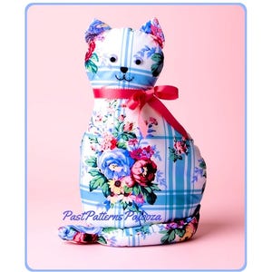 May include: A stuffed cat toy made of white fabric with a blue plaid pattern and a colorful floral print featuring red, blue, and yellow flowers. The cat has googly eyes, a black stitched nose, and a bright pink ribbon tied around its neck.