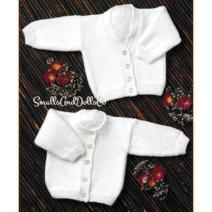 May include: Two white knitted baby cardigans with button closures. The cardigans have long sleeves and a V-neck and round neck. The image includes the text "Smalls And Dolls Co" and is decorated with small red and yellow flowers.