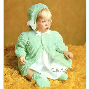 以下が含まれることがあります： 緑色のニットの服を着た赤ちゃんが、それに合わせたボンネットをかぶっています。服はボタン付きのカーディガン、ワンピース、ブーティで構成されています。赤ちゃんは黄色のふわふわした表面に座っています。画像の右下隅には「Smalls And Dolls Co」というテキストが表示されています。