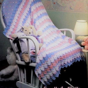 May include: A crocheted baby blanket in shades of pink, blue and white with a chevron pattern. The blanket is draped over a rocking chair with a stuffed animal sitting on it.