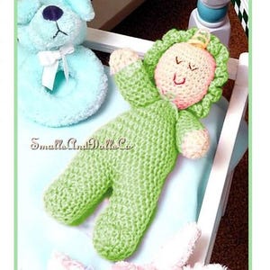 May include: A handmade green crochet baby doll with a smiling face and a floral headpiece. The doll is lying on a light blue surface next to a blue plush dog toy and a white crib. The text "Smalls And Dolls Co." is visible.
