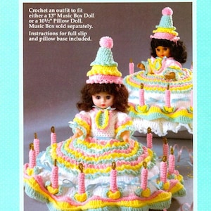 Könnte beinhalten: Häkelanleitung für ein Geburtstags-Puppenkleid. Die Puppe trägt ein weißes Kleid mit rosa, gelben und grünen Rüschen. Die Puppe sitzt auf einem Kuchen-förmigen Sockel mit Kerzen.