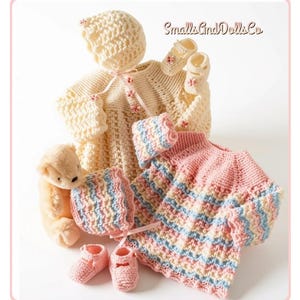 May include: A set of two crocheted baby outfits with matching bonnets and booties. One outfit is cream with pink flower accents, the other is striped in pink, blue, and cream. A small plush bear is included. The text SmallsAndDollsCo is visible.
