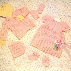 May include: A pink knitted baby outfit set, including a dress, cardigan, hat, and booties. The set is decorated with a delicate lace pattern and features a ribbon tied around the dress.