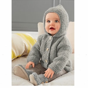 May include: A grey knitted hooded cardigan with button fastenings, worn by a baby sitting on a white surface. The baby is wearing grey knitted booties.