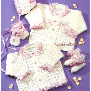 May include: A knitted baby set in cream with pink trim, including a bonnet, two sweaters, and booties. The set features a delicate openwork design and is accented with pink ribbon and buttons. The text "Small and Dolls Co" is visible.