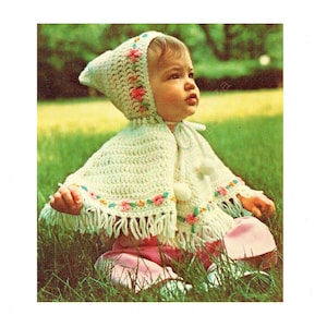May include: A baby wearing a crocheted white hooded cape with a floral trim and fringe. The cape has a pom-pom on the front. The baby is sitting on a grassy lawn.