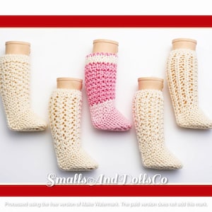 May include: Five knitted doll socks displayed on mannequin feet. Four socks are cream-coloured with varied textured knit patterns, and one central sock features a pink and white colour-blocked design. The text SmallsAndDollsCo is visible at the bottom.